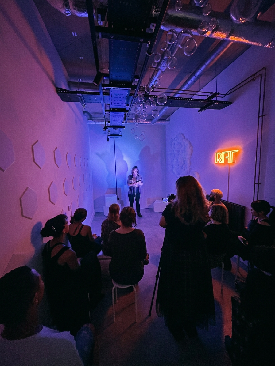 Audience watching a reading beneath the polyfill cloud wall and RIFT neon